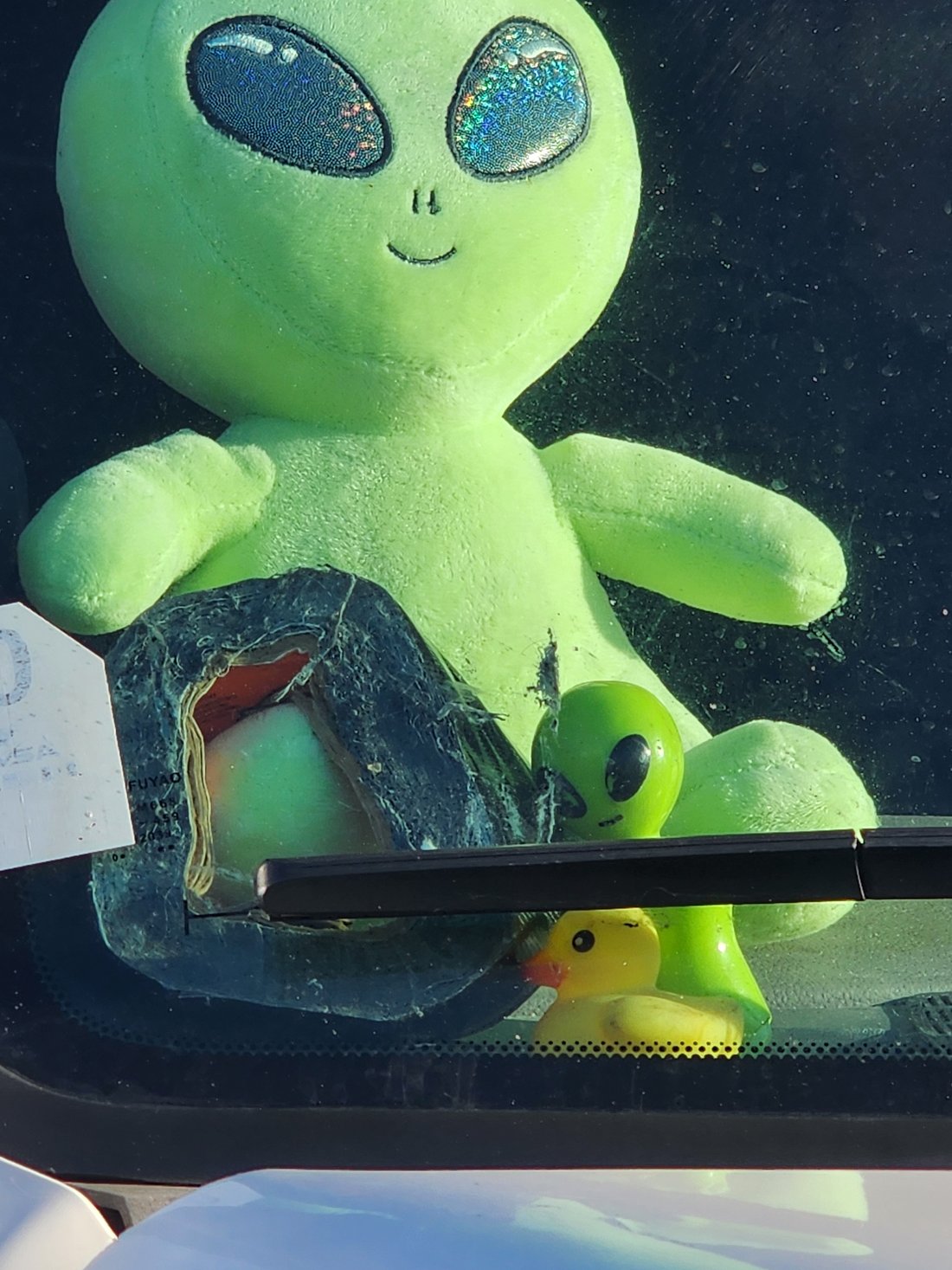 The green alien plush from another angle, with a small rubber duck beside it on the dashboard